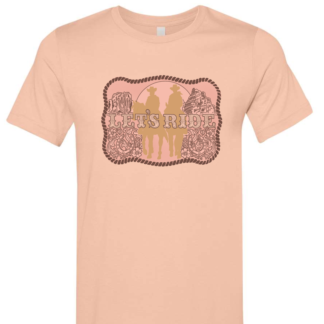 A peach colored tee with a graphic of a belt buckle. Belt buckle is a pink color with a dark brown rope border and brown western filigree on the sides with two cowboys on horses in light tan in the middle of the belt buckle and the words Let's Ride in silver across the width of the belt buckle.