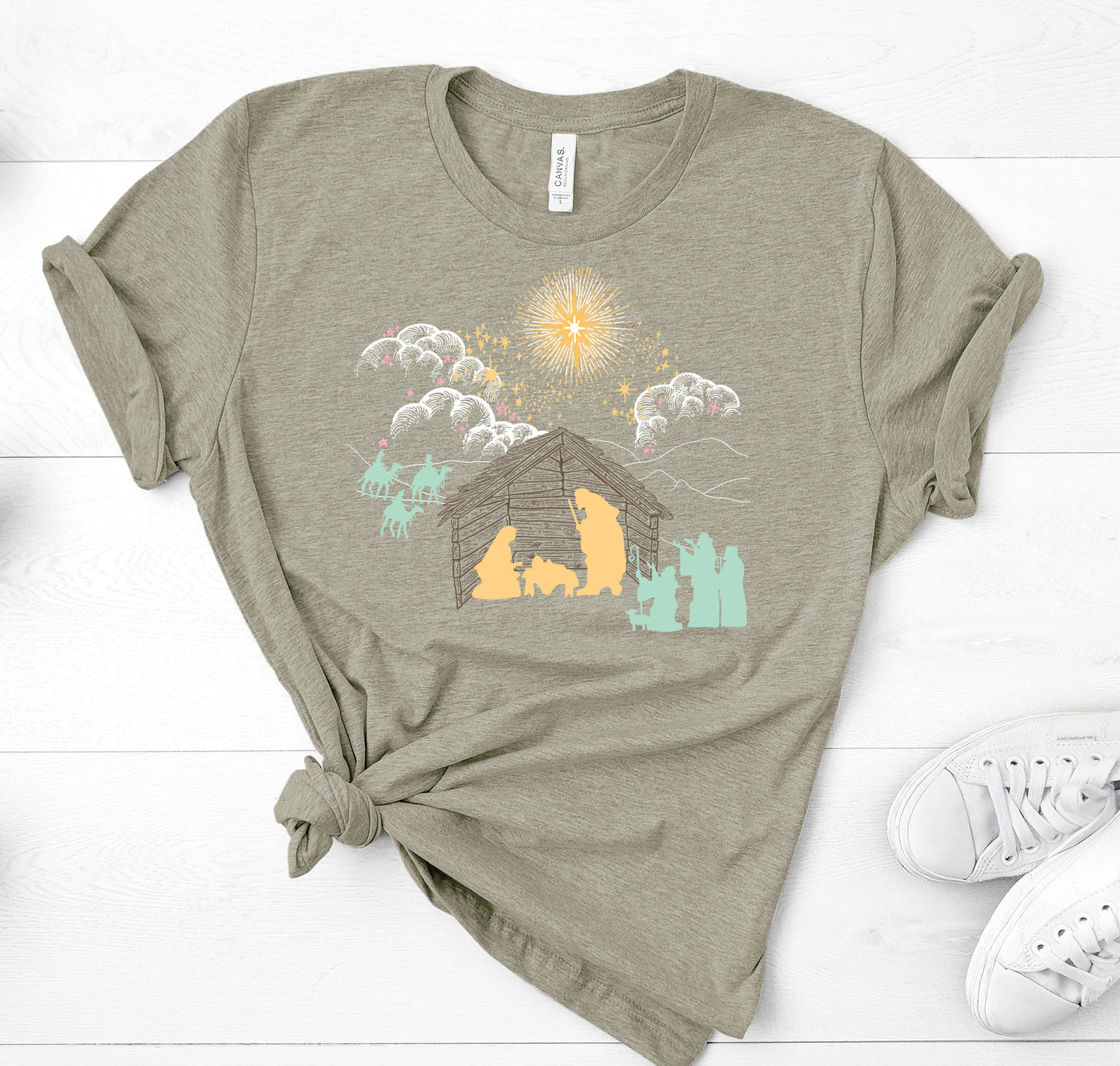 Heather Olive Green T Shirt depicting a nativity scene. White clouds Gold Stars. Yellow Mary Joseph and baby Jesus silhouette, all other silhouettes are light teal