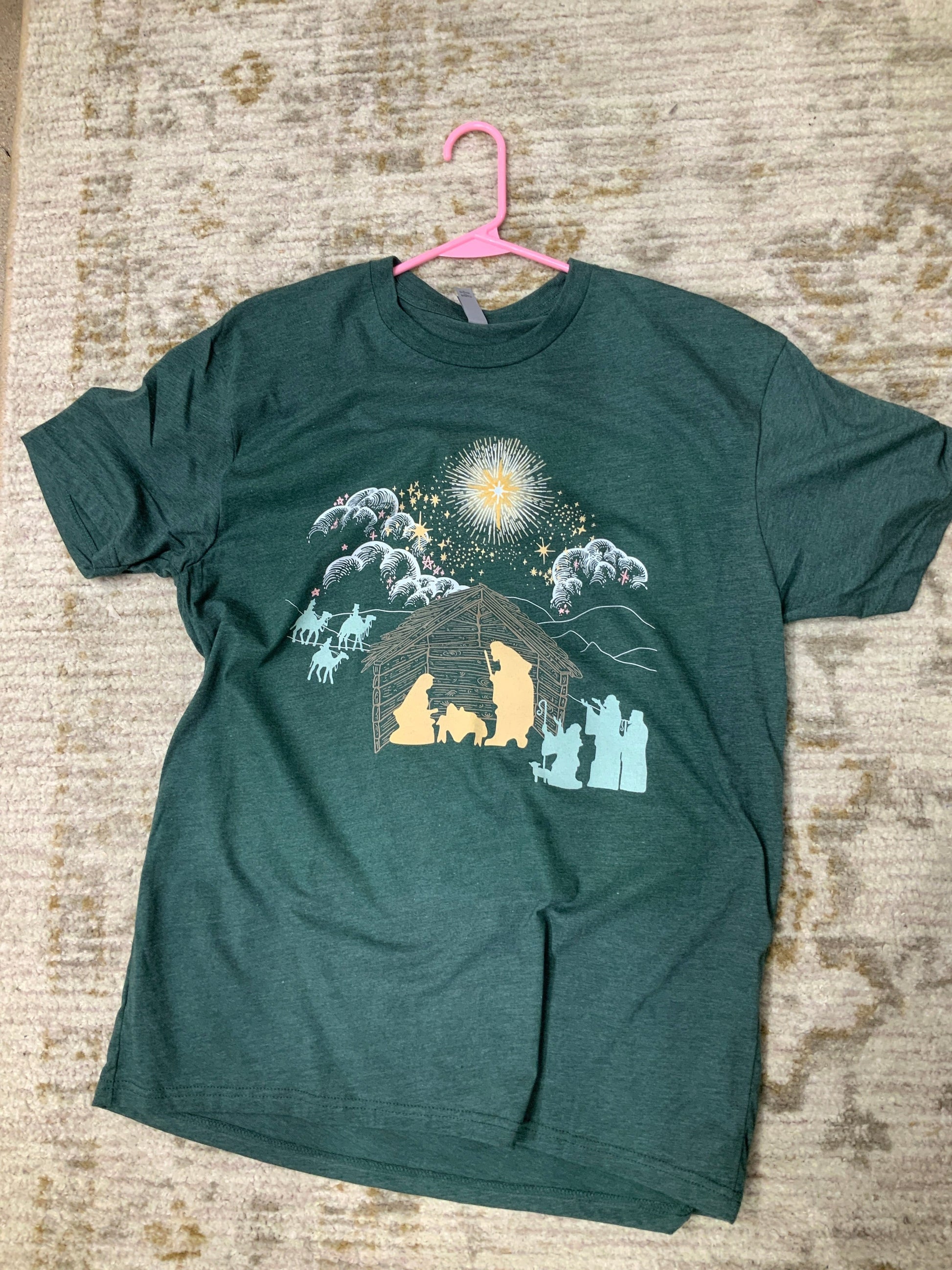 Heather Forest Green T Shirt depicting a nativity scene. White clouds Gold Stars. Yellow Mary Joseph and baby Jesus silhouette, all other silhouettes are light teal